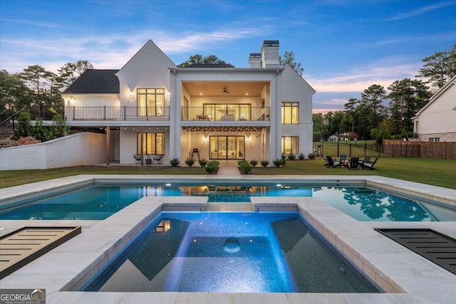 back house at dusk featuring a pool with hot tub, a balcony, and a yard