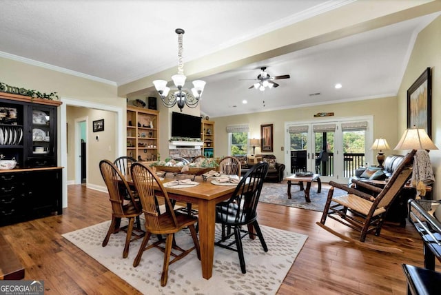 dining area featuring ceiling fan with notable chandelier, wood-type flooring, ornamental molding, and a stone fireplace