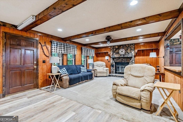 living room with beam ceiling, ceiling fan, a stone fireplace, light hardwood / wood-style flooring, and wooden walls