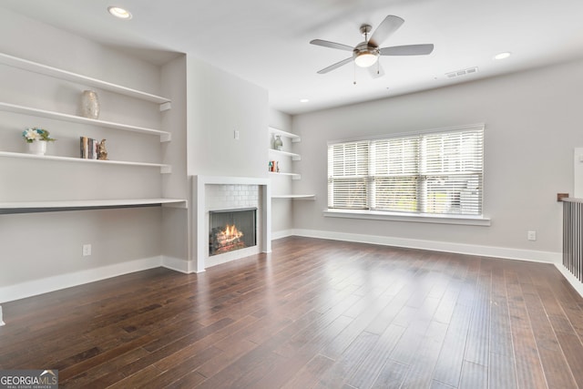 unfurnished living room with ceiling fan, a tiled fireplace, dark wood-type flooring, and built in shelves