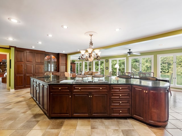 kitchen with pendant lighting, dark stone counters, a large island with sink, ceiling fan with notable chandelier, and stainless steel cooktop