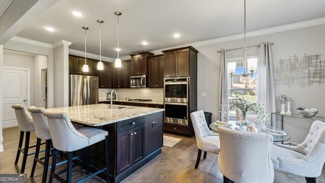 kitchen with wood-type flooring, a center island with sink, appliances with stainless steel finishes, dark brown cabinetry, and decorative light fixtures