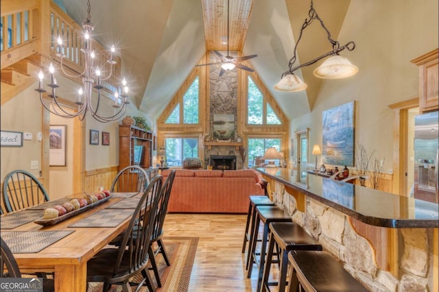 dining area with wood walls, a stone fireplace, ceiling fan with notable chandelier, a towering ceiling, and light hardwood / wood-style floors