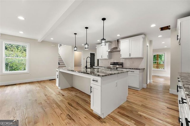 kitchen with dark stone countertops, a center island with sink, wall chimney range hood, white cabinetry, and stainless steel range with electric cooktop