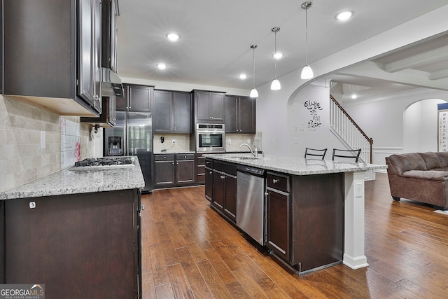 kitchen with appliances with stainless steel finishes, dark wood-type flooring, an island with sink, pendant lighting, and dark brown cabinetry