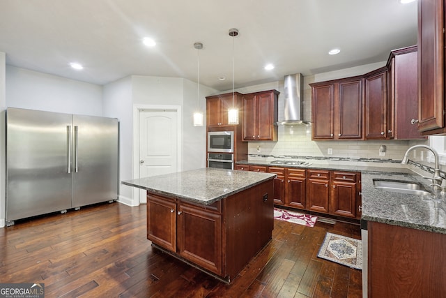 kitchen with sink, a kitchen island, dark wood-type flooring, wall chimney range hood, and appliances with stainless steel finishes