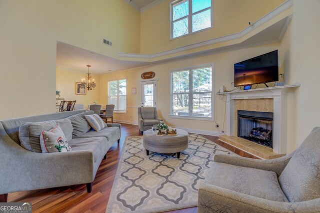 living room with wood-type flooring, a tiled fireplace, an inviting chandelier, ornamental molding, and a high ceiling