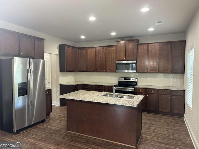 kitchen with light stone counters, dark hardwood / wood-style floors, stainless steel appliances, a center island with sink, and dark brown cabinetry