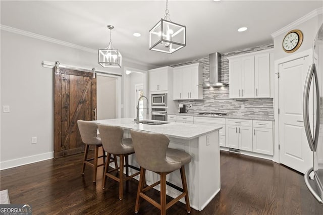 kitchen featuring wall chimney range hood, pendant lighting, an island with sink, white cabinets, and appliances with stainless steel finishes