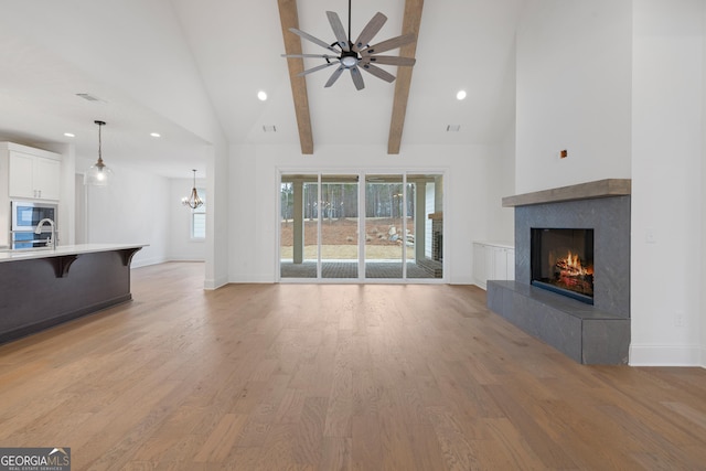 unfurnished living room with high vaulted ceiling, light hardwood / wood-style flooring, beam ceiling, a tiled fireplace, and ceiling fan with notable chandelier