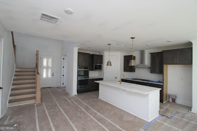 kitchen featuring cooktop, hanging light fixtures, double oven, wall chimney range hood, and a kitchen island with sink