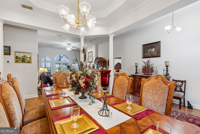 dining area with a raised ceiling, ornamental molding, wood-type flooring, ceiling fan with notable chandelier, and ornate columns