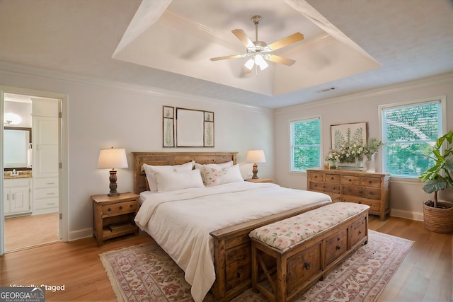 bedroom with ceiling fan, ensuite bath, a tray ceiling, light hardwood / wood-style flooring, and ornamental molding