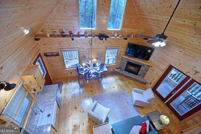 living room with high vaulted ceiling, ceiling fan with notable chandelier, a stone fireplace, hardwood / wood-style flooring, and wooden walls