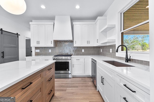 kitchen with open shelves, stainless steel appliances, a barn door, a sink, and premium range hood