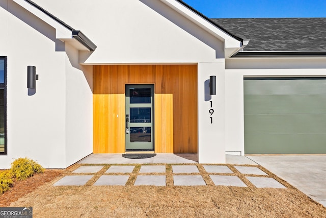 view of exterior entry featuring an attached garage, roof with shingles, and stucco siding