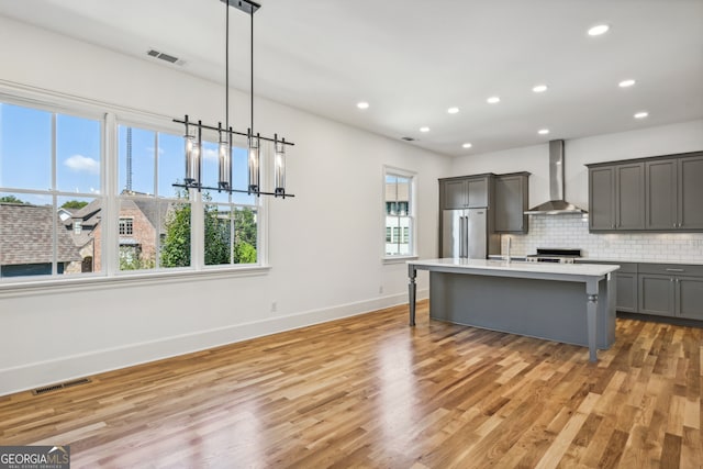 kitchen with pendant lighting, high quality fridge, a center island with sink, wall chimney exhaust hood, and light hardwood / wood-style flooring