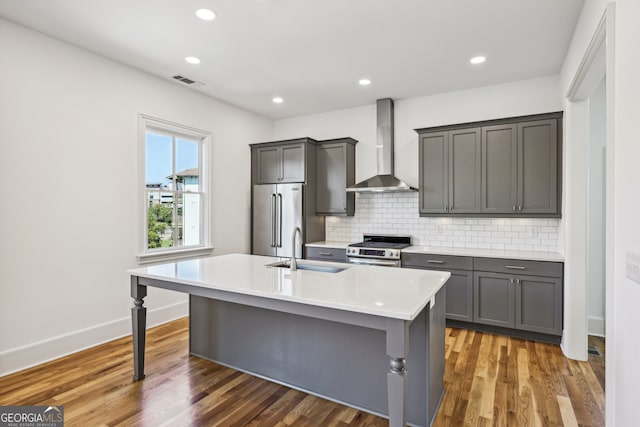 kitchen with an island with sink, sink, wall chimney range hood, appliances with stainless steel finishes, and dark hardwood / wood-style flooring