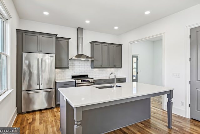 kitchen featuring sink, a center island with sink, wall chimney range hood, hardwood / wood-style flooring, and appliances with stainless steel finishes