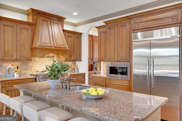 kitchen with light stone countertops, custom exhaust hood, a center island with sink, built in appliances, and a kitchen bar