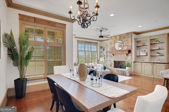 dining room with ceiling fan with notable chandelier, ornamental molding, a stone fireplace, and light hardwood / wood-style floors