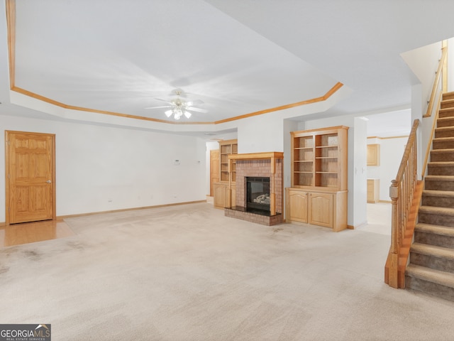 unfurnished living room featuring a brick fireplace, ceiling fan, light colored carpet, a raised ceiling, and ornamental molding