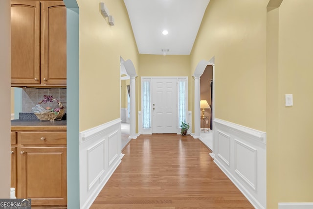 foyer featuring arched walkways, light wood-style flooring, a decorative wall, wainscoting, and decorative columns