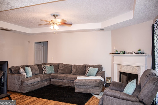 living room featuring ceiling fan, wood-type flooring, a textured ceiling, a tray ceiling, and a fireplace