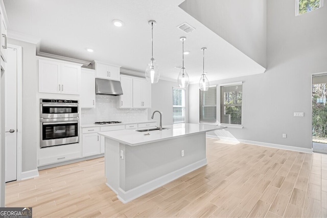 kitchen with a center island with sink, stainless steel double oven, hanging light fixtures, sink, and white cabinets