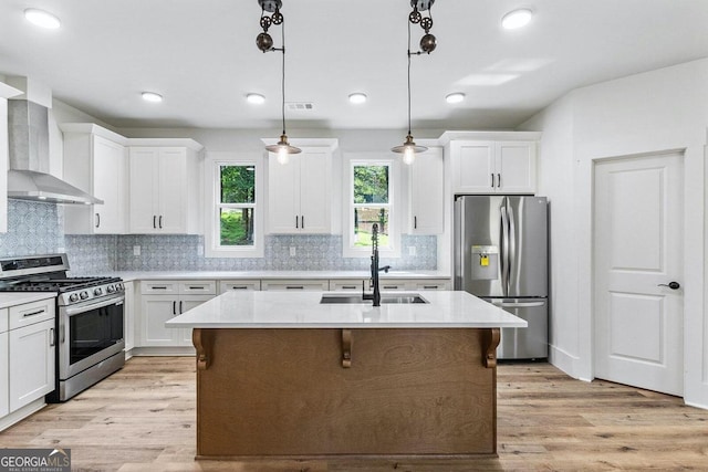 kitchen with decorative light fixtures, a kitchen island with sink, wall chimney exhaust hood, stainless steel appliances, and light hardwood / wood-style floors