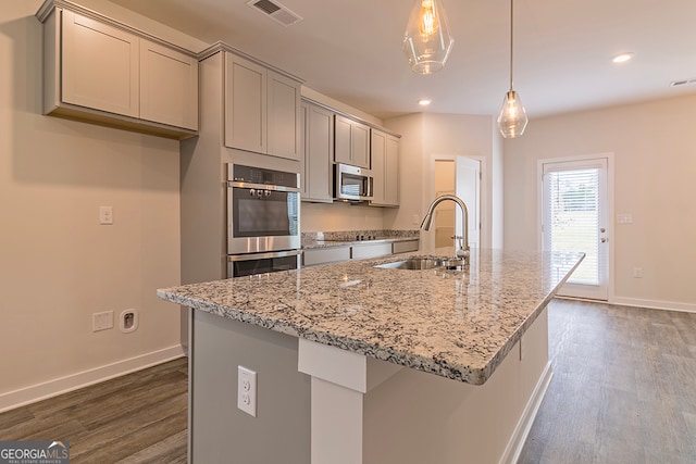 kitchen featuring a center island with sink, sink, light stone counters, stainless steel appliances, and hanging light fixtures