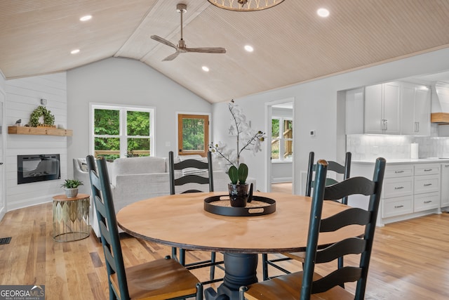 dining area with ceiling fan, wood walls, a large fireplace, light wood-type flooring, and vaulted ceiling