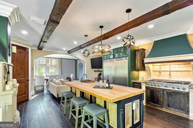 kitchen with beam ceiling, dark hardwood / wood-style flooring, appliances with stainless steel finishes, a kitchen island with sink, and butcher block countertops