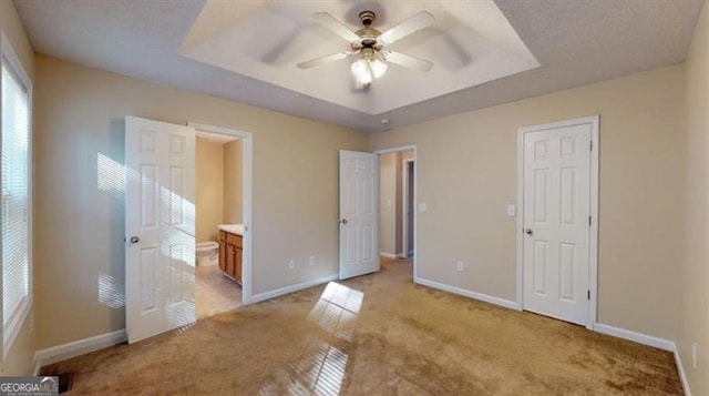 unfurnished bedroom with a raised ceiling, light carpet, ensuite bathroom, and ceiling fan