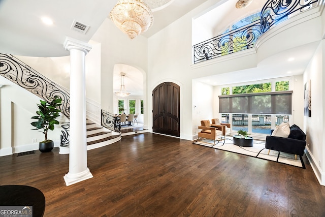 entryway featuring a towering ceiling, ornate columns, dark wood-type flooring, and an inviting chandelier