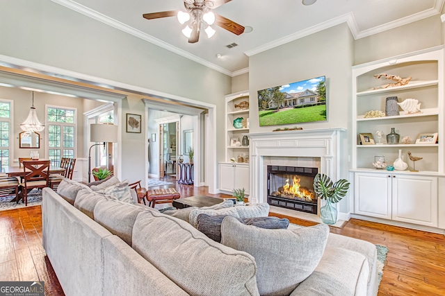 living room with light hardwood / wood-style floors, built in shelves, ceiling fan, a tiled fireplace, and ornamental molding