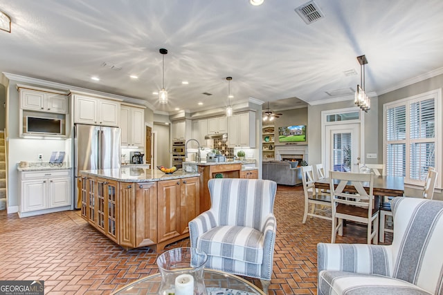 kitchen with an island with sink, light stone counters, stainless steel appliances, hanging light fixtures, and ornamental molding