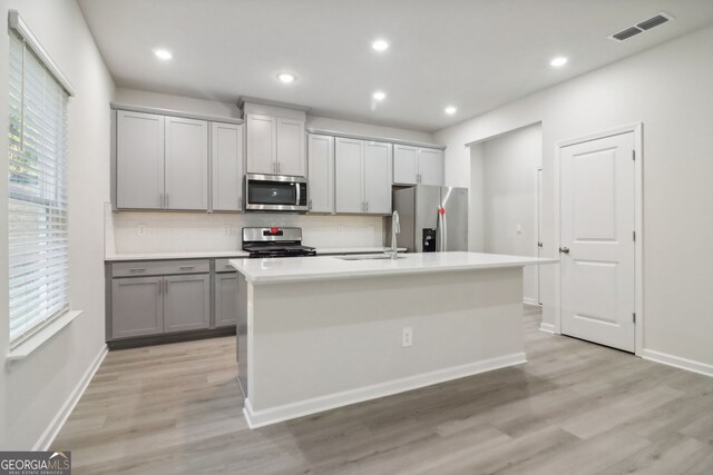 kitchen with light hardwood / wood-style flooring, stainless steel appliances, a center island with sink, gray cabinets, and tasteful backsplash