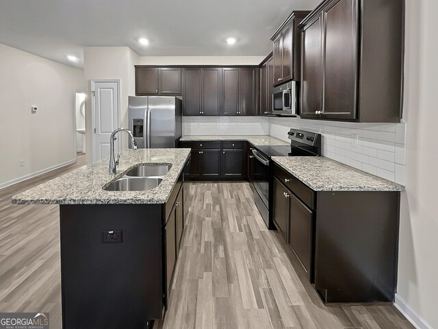 kitchen with light wood-type flooring, light stone counters, sink, appliances with stainless steel finishes, and a center island with sink