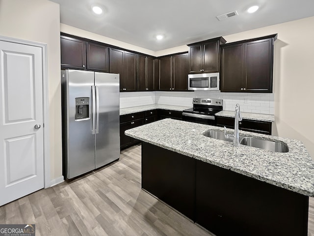 kitchen with a kitchen island with sink, light wood-type flooring, sink, light stone countertops, and appliances with stainless steel finishes