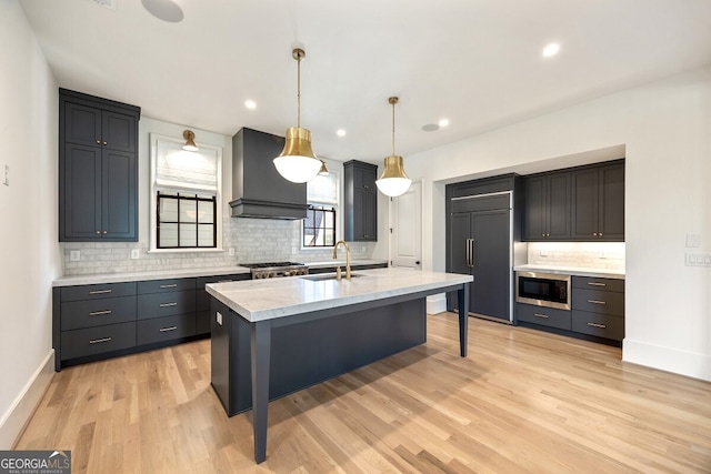 kitchen with custom exhaust hood, decorative light fixtures, a kitchen island with sink, light hardwood / wood-style flooring, and built in appliances