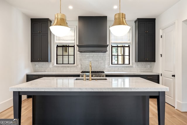 kitchen with a center island with sink, light wood-type flooring, light stone countertops, and wall chimney range hood