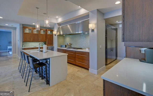 kitchen featuring sink, wall chimney exhaust hood, an island with sink, hanging light fixtures, and decorative backsplash