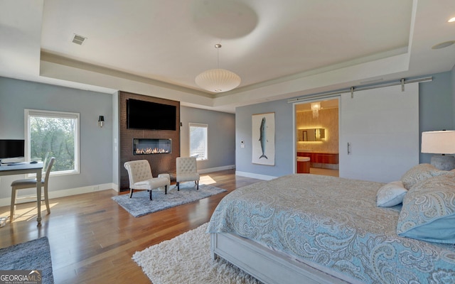 bedroom with ensuite bathroom, a large fireplace, a tray ceiling, hardwood / wood-style floors, and a barn door