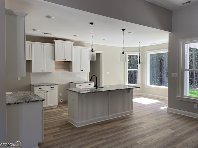 kitchen featuring wood-type flooring, hanging light fixtures, white cabinetry, a center island with sink, and stone countertops