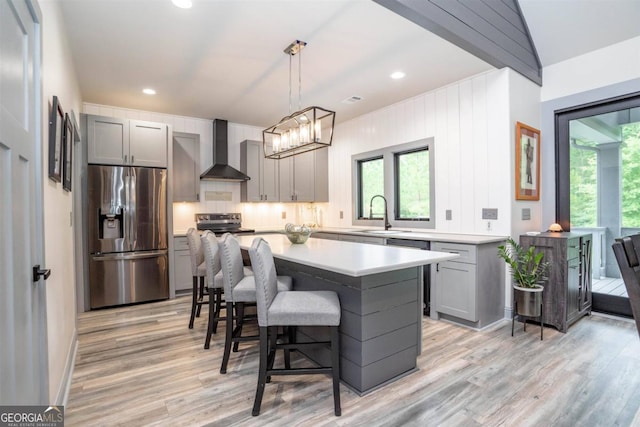 kitchen with hanging light fixtures, gray cabinetry, wall chimney exhaust hood, stainless steel appliances, and light hardwood / wood-style floors