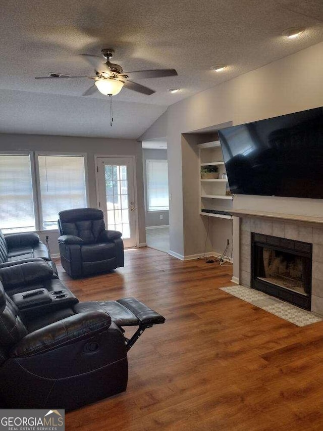 living room featuring ceiling fan, wood-type flooring, a tiled fireplace, vaulted ceiling, and a textured ceiling