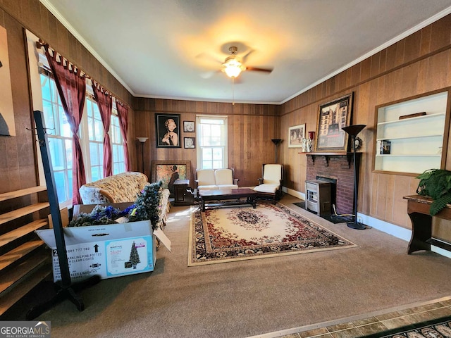 sitting room with wooden walls, ceiling fan, a wood stove, carpet floors, and ornamental molding