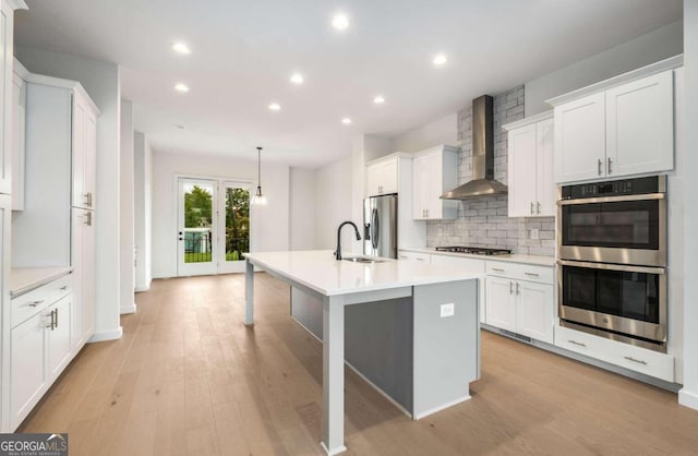 kitchen featuring wall chimney range hood, light hardwood / wood-style floors, stainless steel appliances, hanging light fixtures, and white cabinetry