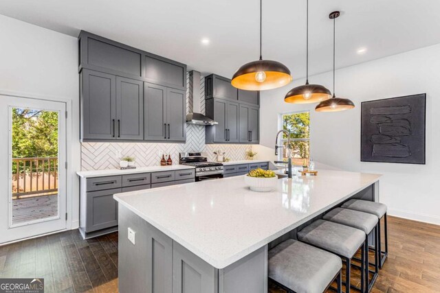 kitchen with gray cabinetry, stainless steel range with gas stovetop, dark hardwood / wood-style flooring, wall chimney range hood, and hanging light fixtures